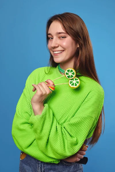 照片上漂亮而快乐的女孩 身穿鲜艳的绿色毛衣 手里拿着两片棒棒糖 一边微笑 一边看着相机 与蓝色背景隔离 — 图库照片