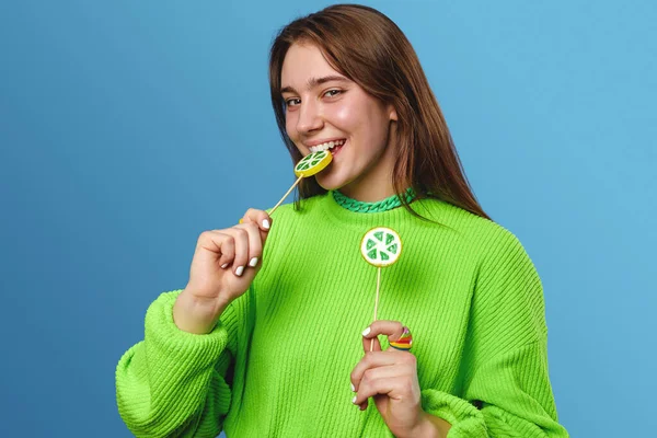 快乐的年轻女士 棕色的头发 穿着绿色的毛衣 快乐地微笑着 做着滑稽的脸 同时在蓝色背景下吃着美味的棒棒糖 — 图库照片