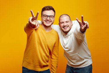 Birbirlerini kucaklayan ve kameraya bakarken barış jesti yapan iki çekici genç adam, sarı turuncu stüdyo arka planında izole bir şekilde duruyorlar. Gerçek arkadaşlık kavramı