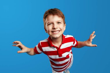 Kırmızı çizgili polo tişörtlü, kollarını kaldırıp eğlenen, mavi arka planda tek başına duran neşeli çocuk portresi. Çocuk stüdyo resmi.