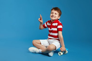 Heyecanlı kırmızı çizgili polo tişörtlü çocuk mavi arka planda modern kaykayın üzerinde otururken gülümsüyor ve barış jesti gösteriyor.