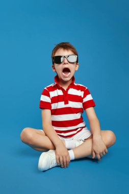Kırmızı çizgili tişört giyen çılgın bir çocuğun dikey fotoğrafı stüdyo zemininde mavi arka planda bacak bacak bacak üstüne atmış otururken yüksek sesle bağırıyor..