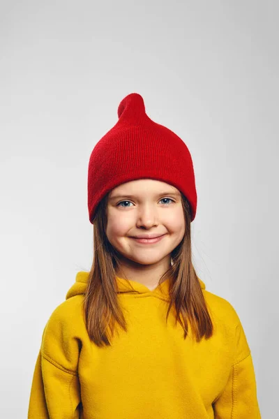 ホワイトバックで孤立したイエローフードと赤い帽子を身に着け 楽しい笑顔で愛らしいヒップスターの子供の垂直近の肖像 — ストック写真