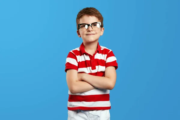 赤いストライプのシャツと神経質な眼鏡でポジティブな未就学児の少年は腕を横切って明るい青い背景に対してカメラを見ています — ストック写真