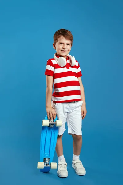 男孩脖子上穿着红色条纹马球衫和耳机 靠在滑板上 对着蓝色背景的相机微笑的全身型时尚男孩的垂直照片 — 图库照片