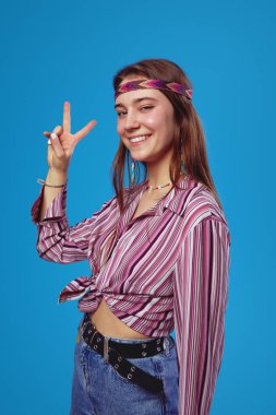 Mavi arka planda izole bir şekilde gülümserken V işareti gösteren pembe tişörtlü çekici genç hippi kızın dikey fotoğrafı. Boho tarzı