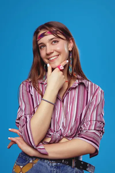 一个穿着粉色衬衫的年轻嬉皮士女人的竖直肖像 手指放在下巴上向上看和思考 隔离在蓝色工作室的背景下 — 图库照片
