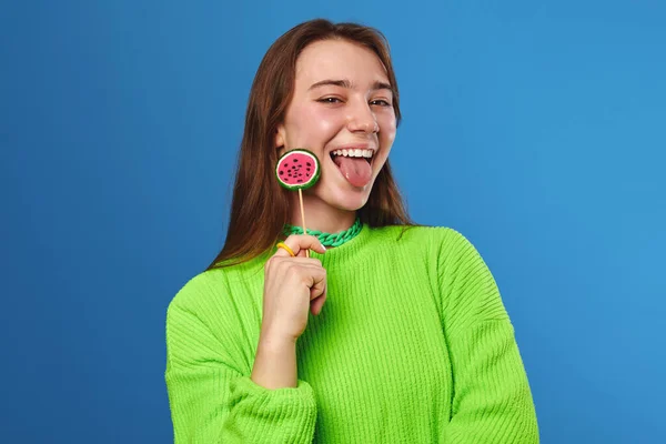 穿着绿色毛衣 嬉闹的少女 嘴上挂着甜瓜棒棒糖 脸上挂着欢乐的表情 与蓝色背景隔离在一起 — 图库照片