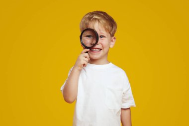 Heyecanlı, beyaz tişört giyen, büyüteçten kafasını çeviren ve sarı stüdyo arka planına karşı durarak gülümseyen meraklı çocuk. Eğitim, ilkokul konsepti