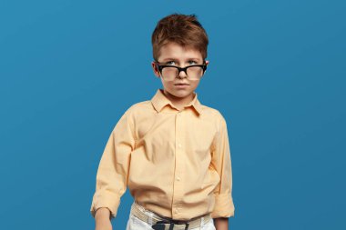 Düşünceli, bej tişörtlü, gözlüklü, bej gömlekli, mavi arka planda tek başına duran küçük bir çocuk..