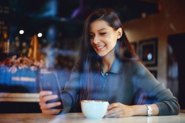 Güzel esmer kadın bir kafede oturuyor ve akıllı telefon kullanıyor. Kahve dükkanında oturan ve cep telefonundan bir şeyler okuyan tatlı bir kız..