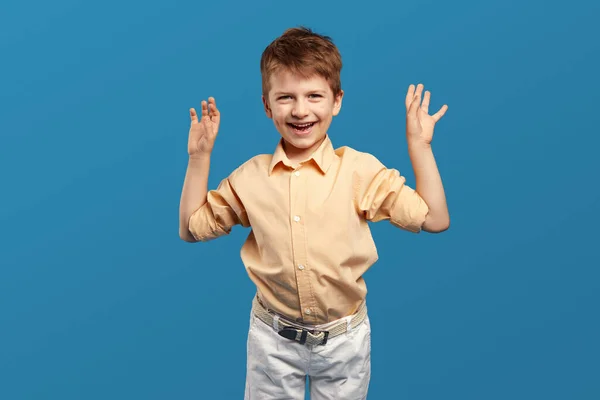 兴奋的英俊男孩穿着米色衬衫 高举双臂庆祝胜利 与蓝色背景隔离 儿童工作室肖像画 — 图库照片