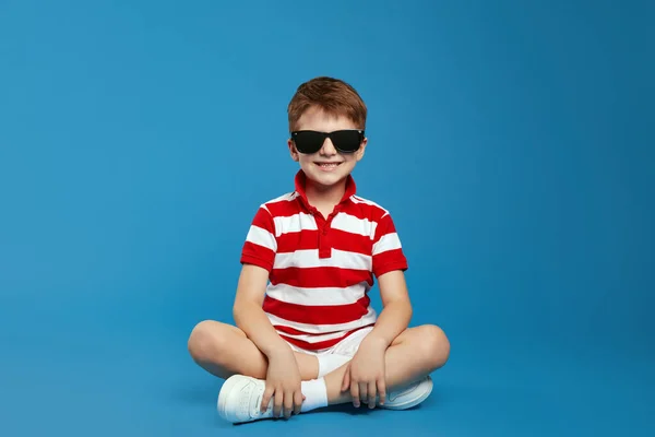 stock image Cool preteen boy in casual outfit and trendy sunglasses feels happy sitting against the blue background of the studio and looking at the camera, portraying an important guy.