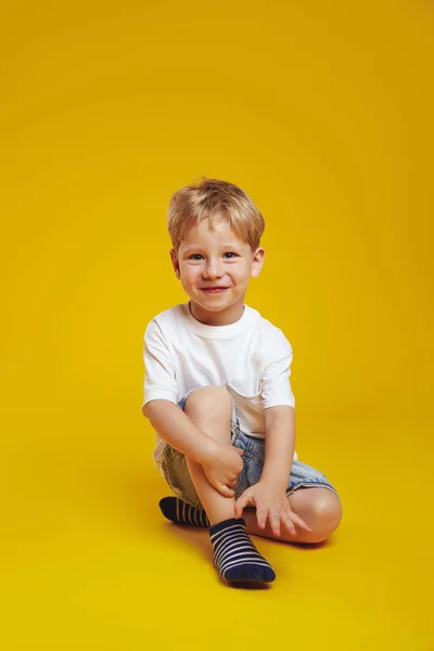 트렌디 티셔츠에 귀여운 소년의 미소와 스튜디오 바닥에 앉아있는 카메라를보고 노란색 스톡 이미지