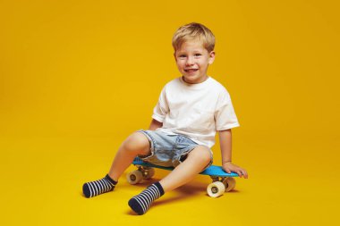 Beyaz tişörtlü ve kot şortlu havalı küçük çocuk mavi modern kaykayın üzerinde otururken ve kameraya gülümseyerek bakarken mutlu oluyor. Stüdyoda poz veren sevimli çocuk, sarı arka planda izole edilmiş.