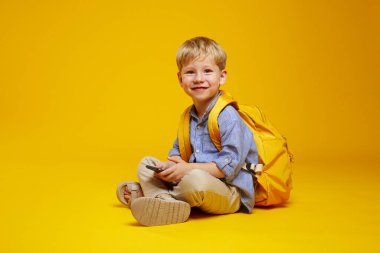 Mavi tişörtlü ve sarı sırt çantalı heyecanlı bir öğrenci stüdyo zemininde bacak bacak üstüne atmış, sarı arka planda izole bir şekilde otururken elinde bir telefon tutuyor..