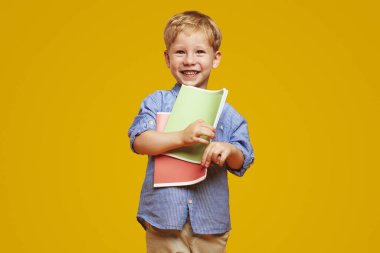 Heyecanlı, mavi tişörtlü, elinde iki defterle gülümseyen, okula hazır, sarı arka planda izole bir çocuk..