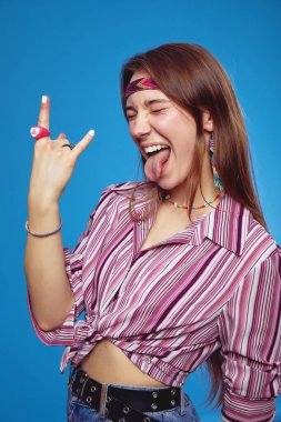 Heyecanlı genç hippi kadının dikey fotoğrafı, elleri havada, dili kapalı, mavi arka planda izole bir şekilde rock n roll hareketi yapıyor.