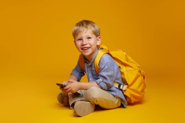 Positive schoolboy with yellow rucksack sitting on studio floor with crossed legs, using mobile phone and looking at camera excited. Isolated over yellow background.