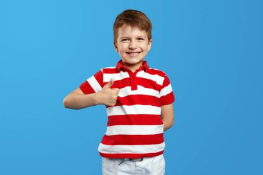 Little preschool boy smiling and wearing red striped polo shirt, posing on blue background while showing thumb up. Portrait studio shot