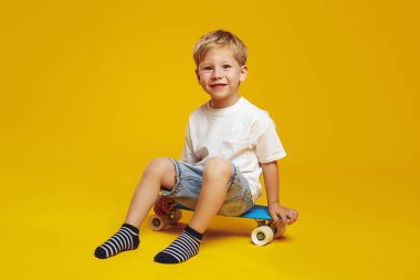 Full length of positive little boy model in stylish outfit smiling for camera and sitting on modern blue skateboard, isolated over yellow background.