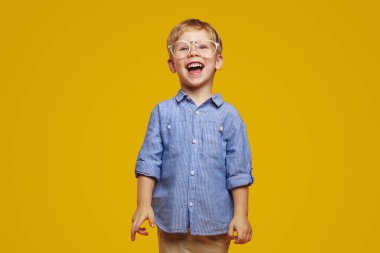 Cheerful funny little kid with blonde hair in eyeglasses, smiling brightly and looking at camera while standing against yellow background.