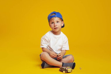Beyaz tişörtlü ve mavi şapkalı küçük bir çocuk, stüdyo zemininde fotoğraf makinesinin yanında otururken, sarı arka planda izole bir şekilde başka tarafa bakıyor..