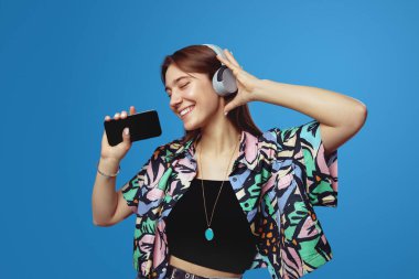 Genç bir kadın mavi arka planda izole edilmiş kulaklıkla en sevdiği müziği dinlerken aktif olarak gözleri kapalı dans ediyor. İnsanlar, yaşam tarzı ve duygu kavramı