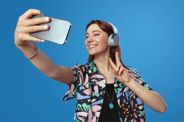 Renkli tişörtlü ve kulaklıklı genç esmer, akıllı telefonlu selfie çekerken barış jesti gösteriyor, mavi arka planda tek başına duruyor. İşaretler, semboller kavramı
