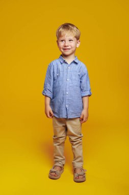 Mavi tişörtlü sevimli sarışın çocuğun dikey fotoğrafı. Sarı arka planda dikilirken gülümsüyor ve başka tarafa bakıyor..
