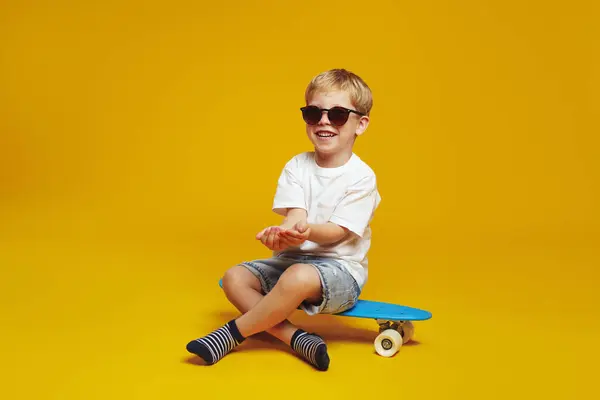 stock image Cool little kid boy wearing casual outfit and sunglasses trying to catch something while sitting on skateboard against yellow studio backdrop