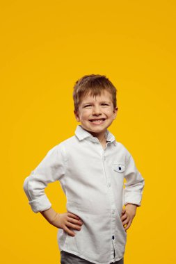 Beyaz gömlekli, kameraya gülümseyen, elleri belinde sarı parlak arka planda gezen küçük çocuğun dikey fotoğrafı.