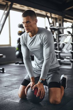 Yakışıklı, kaslı fiziği olan atletik bir adam tıp topuyla yaptığı yoğun çalışma sırasında ara veriyor. Spor kıyafetleri içinde, modern bir spor salonunda odaklanarak ve kararlılıkla diz çöker..