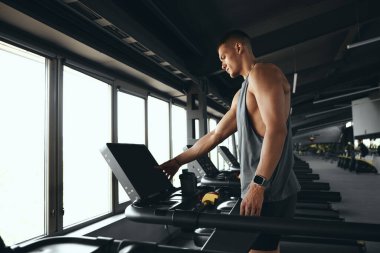 Aktif giyimde atletik adam koşu bandındaki ayarları ayarlıyor, iyi donanımlı spor salonunda kardiyo seansına hazırlanıyor. Sportif erkek koşu pistinde yürüyor, sağlık ve zindelik üzerine çalışıyor.