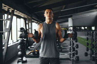 Spor giyim kuşağında vücut geliştiren bir adamın modern spor salonunda kaslarını dambıllarla çalıştırması ön planda. Vücut geliştirme, fitness ve sağlık hizmeti kavramı.