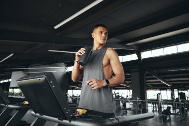 Aktif, gri tişörtlü, şişeden su içen ve koşu bandında dikilen çekici genç bir adam. Spor seansından sonra susuz kalmayan genç bir adam. Spor çalıştırma konsepti.