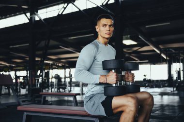 Spor salonunda halterleri tutan güçlü ve formda bir adam. Güç antrenmanına hazır. Doğal ışık ve ekipmanlarla dolu modern bir fitness ortamında odaklanmış ve kararlı bir adam.