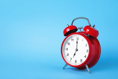 Kırmızı bir retro çalar saat, zaman yönetimi ve dakikliği sembolize eden canlı bir mavi arkaplan karşısında duruyor. Saat 12 'ye ayarlandı, bu da aciliyet ve dikkat hissi uyandırıyor..