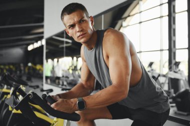 Spor kıyafetleri içinde spor salonunda spor salonunda spor bisikletle spor yapan ciddi bir formda adam. Sporcu erkek egzersiz bisikletinde antrenman yapıyor ve yan bakıyor. Genç adam modern spor salonundaki bir sınıfta çalışıyor..