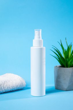 Parlak beyaz bir sprey şişesi, dokulu bir havlu ve saksı bitkisinin yanında duruyor. Sakinleştirici mavi bir zemin üzerine kurulmuş. Tazelik ve sadelik hissi yaratıyor..