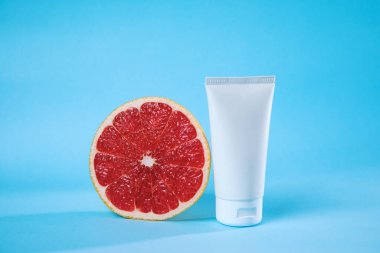 Taze bir greyfurt, canlı bir mavi arkaplan karşısında beyaz bir cilt tüpünün yanında oturur ve sağlık odaklı ve canlandırıcı bir görsel tema yaratır..