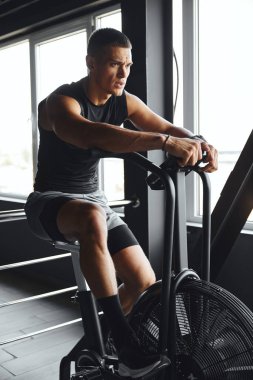 Spor giyim sektöründe yakışıklı atletik adam hava bisikleti üzerinde sıkı çalışıyor, modern bir spor salonunda kardiyo çalışması yapıyor. Odaklanması ve yoğunluğu sağlıklı bir yaşam tarzına ve fitness hedeflerine olan bağlılığını gösteriyor..