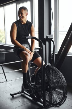 Modern spor salonunda kardiyo çalışması için hava bisikleti kullanan çekici bir fitness oyuncusunun dikey fotoğrafı. Güç eğitimi, disiplin ve iş başındaki yaşam tarzı.