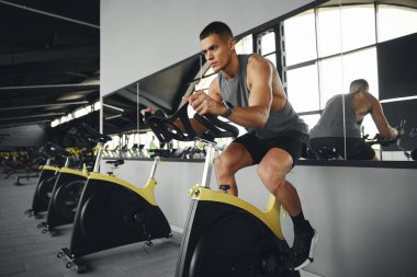 Spor kıyafetleri içinde konsantre olmuş formda bir adam bisiklet üstünde kalori yakıyor. Durağan bisiklete binen yakışıklı sporcu. Spor ve sağlık hizmetleri kavramı.