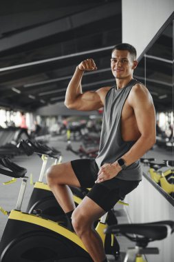 Geniş, modern spor salonunda bisiklet makinesinde otururken spor kıyafetli, gülümseyen ve pazılarını gösteren neşeli genç adamın dikey fotoğrafı..