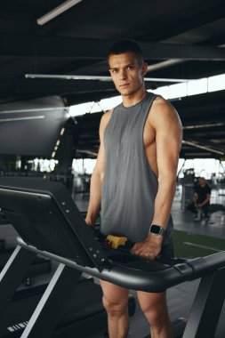 Spor giyimli, dikey bir erkek sporcunun koşu bandında durup iyi donanımlı spor salonunda kardiyo seansına hazırlandığı sahne. Zinde adam ayakta durup kameraya doğru bakıyor, sağlık ve zindelik üzerine çalışıyor..