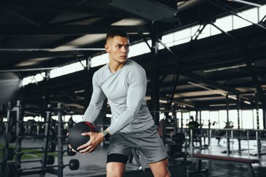 İyi yapılı bir erkeğin spor salonundaki bir ilaç topuyla antrenman yaparken çekilmiş yatay fotoğrafı. Enerjik adam güç ve direnç oluşturmaya odaklanır, öz disiplin değerlerini yansıtır.