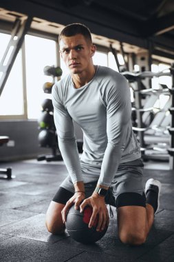 Sporcu kıyafetli yakışıklı genç adam spor salonunda sağlık topuyla antrenman yaparken mola veriyor. Odaklanmış ifade, kas yapısı ve doğal ışıklandırmalı modern fitness ortamı.