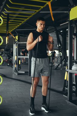 Modern spor salonundaki işlevsel eğitim sırasında spor giyim kuşağında elastik direnç bandıyla egzersiz yapan kendinden emin kaslı bir erkeğin dikey fotoğrafı.
