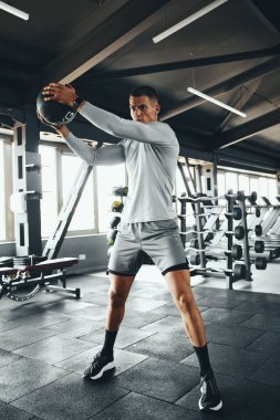 Spor salonunda ilaç topuyla çalışan kaslı bir adamın dikey fotoğrafı. Atletik genç erkek güçlü bir güç rutini içinde, kendini adama ve formda kalma disiplininin temaları vurgulanıyor.